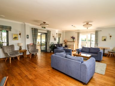 A sitting room with blue sofas, grey armchairs, wooden coffee and side tables, curtains and wooden flooring at Llety Ceiro in Bow Street Wales