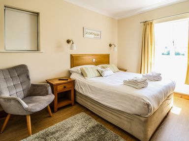 A bedroom with a bed, two bedside tables, a grey armchair, and a window with yellow curtains at Llety Ceiro Bow Street Wales