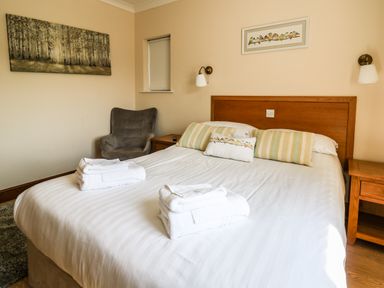 A bedroom with a double bed with pillows and towels two bedside tables a gray armchair and wall art at Llety Ceiro in Bow Street Wales