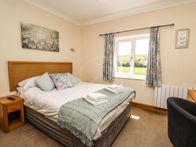 A bedroom with a double bed pillows and folded towels near a window with floral curtains at Llety Ceiro in Bow Street Wales