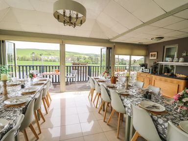 A dining room with two long tables set with plates and glasses facing a balcony with open doors and a view of green fields at Llety Ceiro in Bow Street Wales