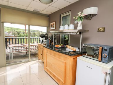A kitchen area with a counter holding cups bread and fruit next to a small fridge and microwave with a sliding door leading to an outdoor dining area at Llety Ceiro in Bow Street Wales