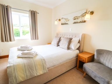 A bedroom with a bed with pillows and towels a window with curtains a wooden nightstand and a gray upholstered chair at Llety Ceiro in Bow Street Wales