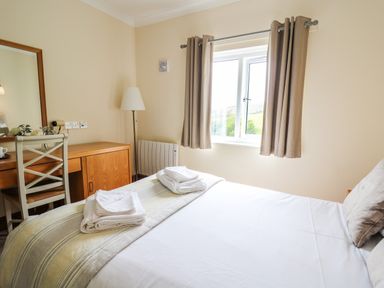 A bedroom with a bed, folded towels, a window with curtains, a lamp, a wooden desk, and a chair at Llety Ceiro in Bow Street Wales