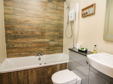 A bathroom with a bathtub shower combination a toilet gray cabinetry and a sink at Llety Ceiro in Bow Street Wales