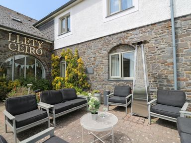 An outdoor seating area with black cushioned chairs and a metal patio heater in front of a stone building at Llety Ceiro Bow Street Wales
