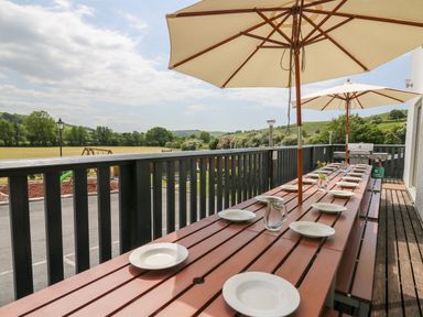 An outdoor dining table with plates glasses jug and umbrellas on a wooden deck overlooking a field with trees at Llety Ceiro in Bow Street Wales