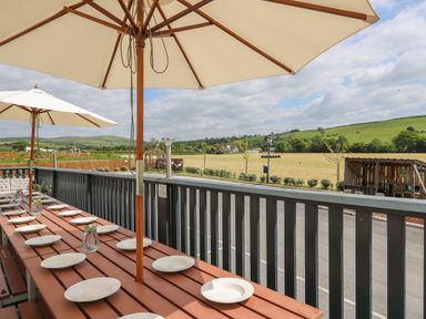 An outdoor dining area with a long wooden table set with plates under large umbrellas overlooking fields at Llety Ceiro in Bow Street Wales