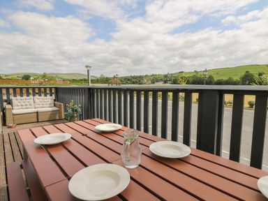 An outdoor dining area with a wooden table set with plates and a glass pitcher and a cushioned bench on a balcony overlooking a rural landscape at Llety Ceiro in Bow Street Wales