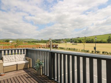A wooden deck with a cushioned wicker sofa and a vase with flowers overlooking a field and hills at Llety Ceiro in Bow Street Wales