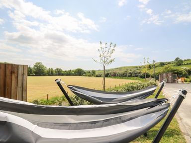 Two striped hammocks on stands near a field with trees and hills in the background at Llety Ceiro in Bow Street Wales