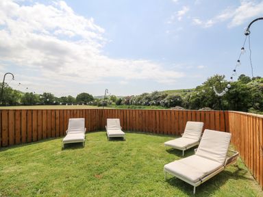 An outdoor grassy area with four lounge chairs enclosed by a wooden fence and string lights at Llety Ceiro in Bow Street Wales