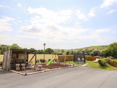 An outdoor play area with a wooden shelter and stools a swing set slide trampoline and lounge chairs at Llety Ceiro in Bow Street Wales