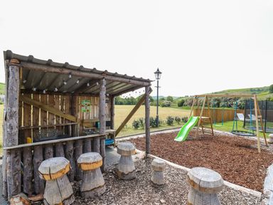 An outdoor play area with wooden stools a swing set with green slide and a small wooden shelter at Llety Ceiro in Bow Street Wales
