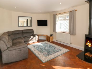 A living room with a gray sectional sofa a blue and white rug a wall-mounted TV a window with gray curtains and a wood-burning stove at Ty Garth in Benllech