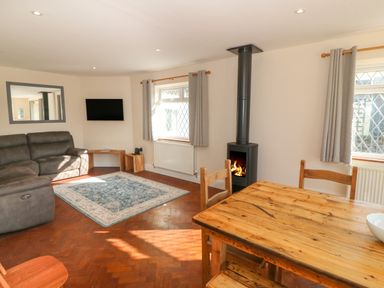 A living room with a gray sofa a wall-mounted TV a wood-burning stove a wooden dining table and chairs at Ty Garth in Benllech