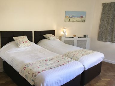 A bedroom with two single beds with patterned pillows and runners a decorative lamp and a painting above a radiator cover at Ty Garth in Benllech
