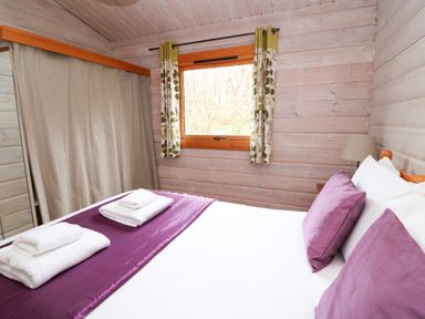 A bedroom with a bed with purple pillows and white towels on a purple runner a window with curtains and wooden walls at Barn Owl Lodge in Rhayader