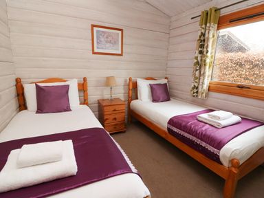 A bedroom with two single beds side by side with purple pillows and throws with a wooden nightstand between at Barn Owl Lodge Rhayader