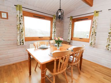 A dining room with a wooden table set for four near two windows with patterned curtains at Barn Owl Lodge in Rhayader