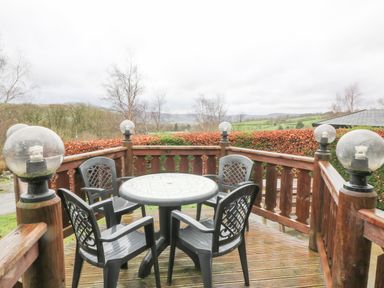 An outdoor wooden deck with a round table and four plastic chairs surrounded by wooden railing and globe lights at Barn Owl Lodge in Rhayader