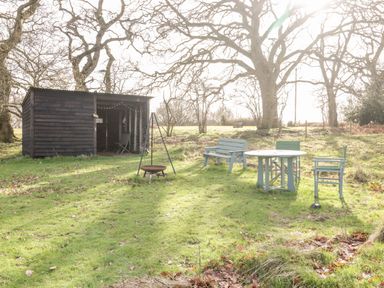 An outdoor area with a wooden shelter chairs a table and a fire pit on grass with trees and a bench in the background at Deer Lodge in Wimborne Minster