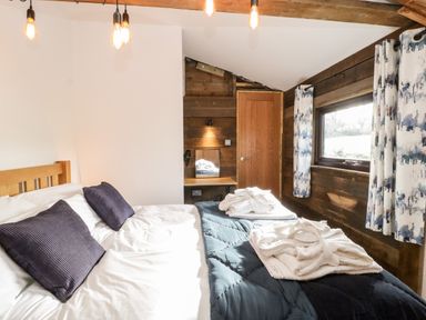 A bedroom with a wooden bed, two purple cushions, a blue quilt, robes on the bed, wooden desk, wooden door, window with patterned curtains at Deer Lodge in Wimborne Minster