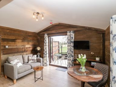 A living room with a gray sofa pillows small wooden table round wooden table with flowers patio doors with curtains and wall mounted tv at Deer Lodge in Wimborne Minster