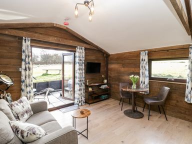 A living room with a gray sofa and small wooden tables next to a glass door and window with patterned curtains at Deer Lodge in Wimborne Minster