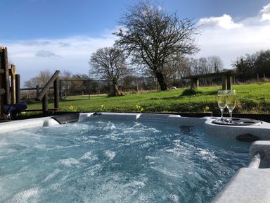 An outdoor hot tub with two glasses of champagne on a tray overlooking a grassy area with trees and a wooden bench at Deer Lodge in Wimborne Minster