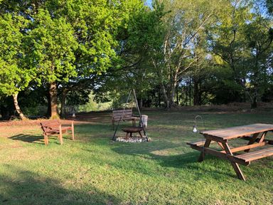 An outdoor seating area with wooden benches and a picnic table around a fire pit in a grassy area with trees at Deer Lodge in Wimborne Minster