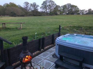 An outdoor patio with a hot tub a fire pit two glasses of wine and a grassy field with trees in the background at Deer Lodge in Wimborne Minster