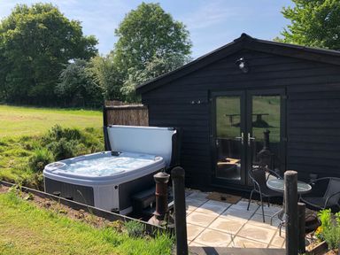 An outdoor patio with a hot tub table and two chairs next to a black wooden building surrounded by grass and trees at Deer Lodge in Wimborne Minster