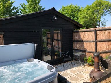 A patio area with a hot tub table with two chairs a chiminea a black wooden building and a fence at Deer Lodge in Wimborne Minster