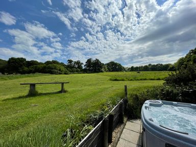 An outdoor area with a hot tub patio bench and a large grassy field with trees under a blue sky with clouds at Deer Lodge in Wimborne Minster