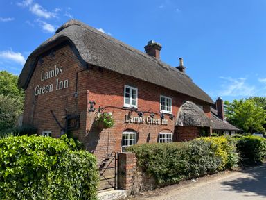 A brick building with a thatched roof and signage for Lambs Green Inn surrounded by green bushes and a clear blue sky at Deer Lodge in Wimborne Minster
