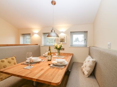 A dining area with a wooden table set with plates glasses cutlery a bottle of wine and flowers with bench seating and three small windows at Larchwood Lodge in Ellon