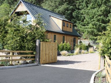 A wooden house with a metal roof behind a wooden gate and surrounded by trees and plants at Larchwood Lodge in Ellon