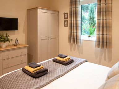 A bedroom with a bed, folded towels on the bed, a wardrobe, a chest of drawers, a wall-mounted television, and a window with curtains at Larchwood Lodge in Ellon