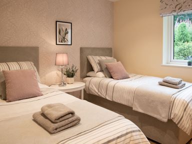A bedroom with two single beds a round white bedside table a lamp framed artwork and a window at Larchwood Lodge Ellon