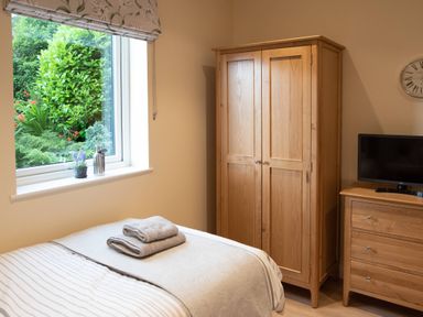 A bedroom with a bed folded towels on it a wooden wardrobe a wooden dresser with a television and a window showing garden plants at Larchwood Lodge in Ellon