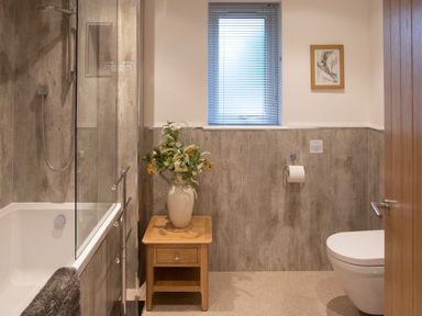A bathroom with a bathtub a wooden side table with a vase of flowers a window with blinds and a wall-mounted toilet at Larchwood Lodge in Ellon