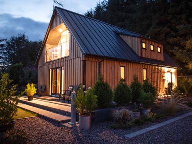 A wooden house with lit windows and a small garden with plants in pots at Larchwood Lodge in Ellon