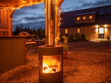 An outdoor area with a burning fire stove string lights a grill and a wooden building in the background at Larchwood Lodge in Ellon