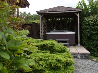 An outdoor patio area with a hot tub under a wooden gazebo surrounded by green bushes at Hillside View in Rhayader