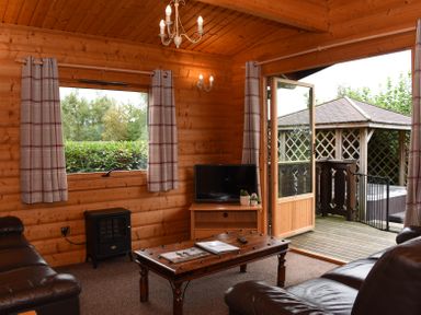 A wooden lounge with leather sofas a wooden coffee table a black heater a television on a stand and a door opening to a deck with a gazebo at Hillside View in Rhayader