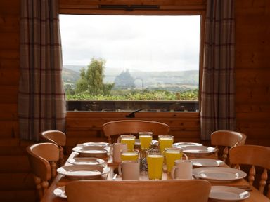 A wooden dining table set with white plates and glasses of orange juice in a room with a large window and plaid curtains at Hillside View in Rhayader
