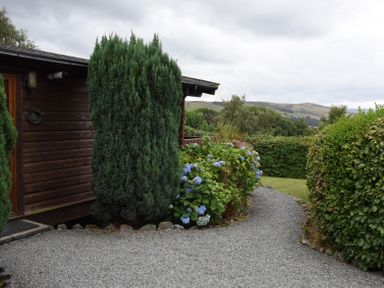 A gravel pathway with bushes and flowers outside a wooden cabin at Hillside View in Rhayader