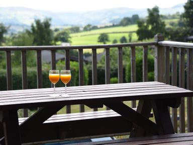Two glasses of orange juice on a wooden picnic table on a balcony with a view of green fields and trees at Sunny View in Rhayader