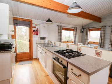 A kitchen with a gas stove oven sink and cabinets next to a dining area with windows and curtains at Snowy Owl Lodge in Rhayader